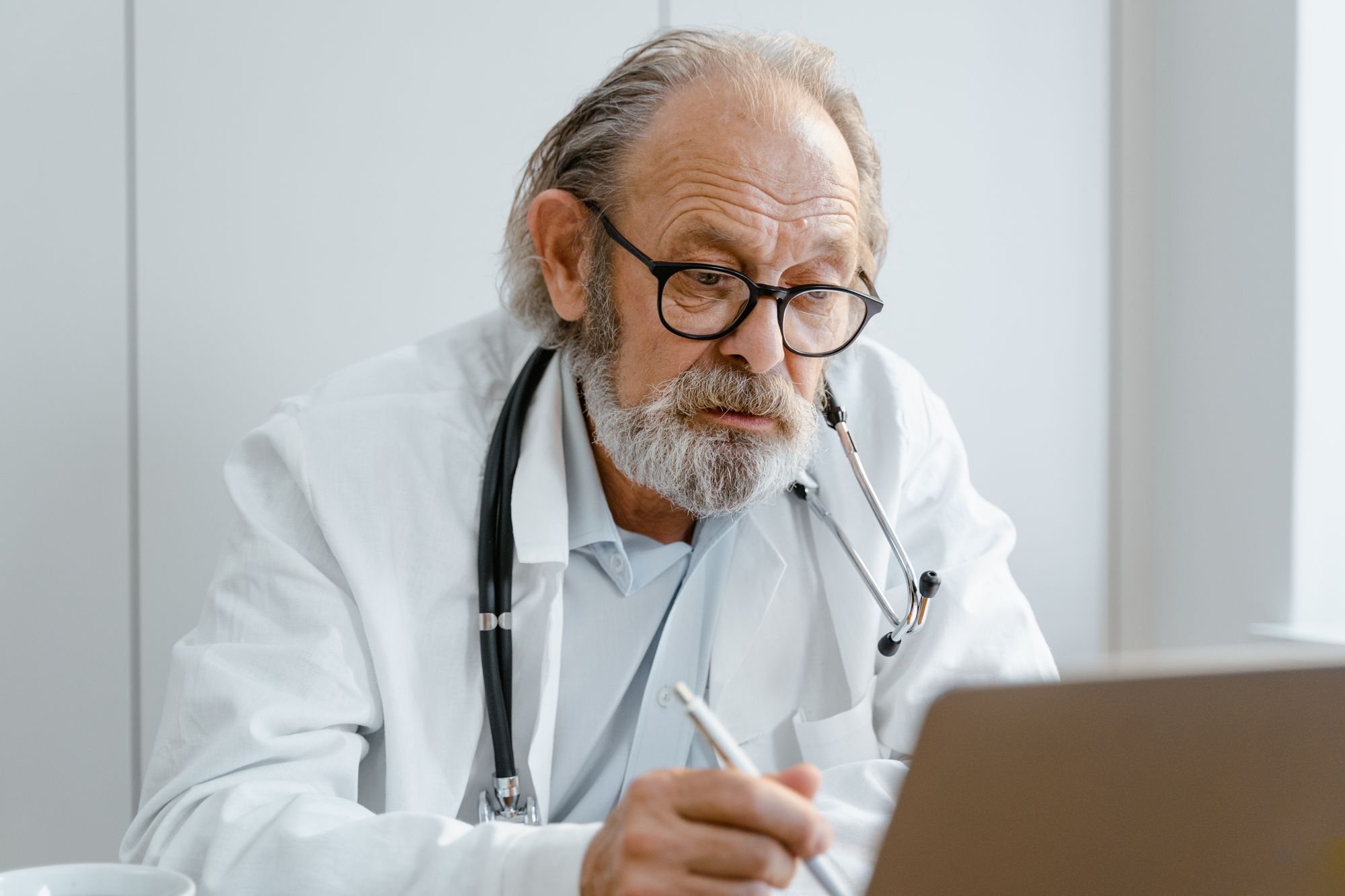 Doctor in a white coat taking notes beside a laptop at his desk