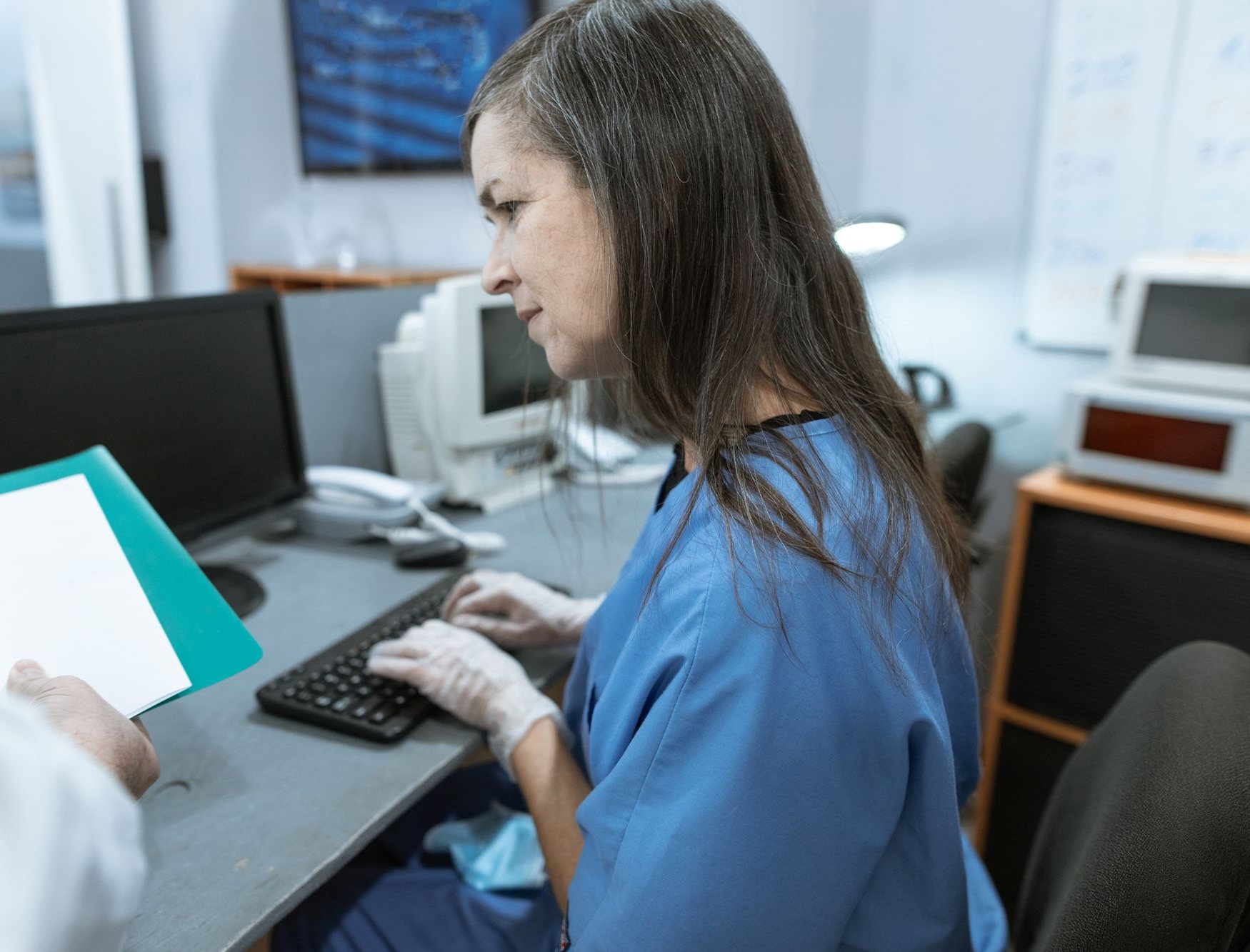 Nurse entering details at a computer as a colleague hands her a clinical document